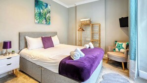 Un dormitorio con cama, mesita de noche, una silla, televisión y un cuadro.