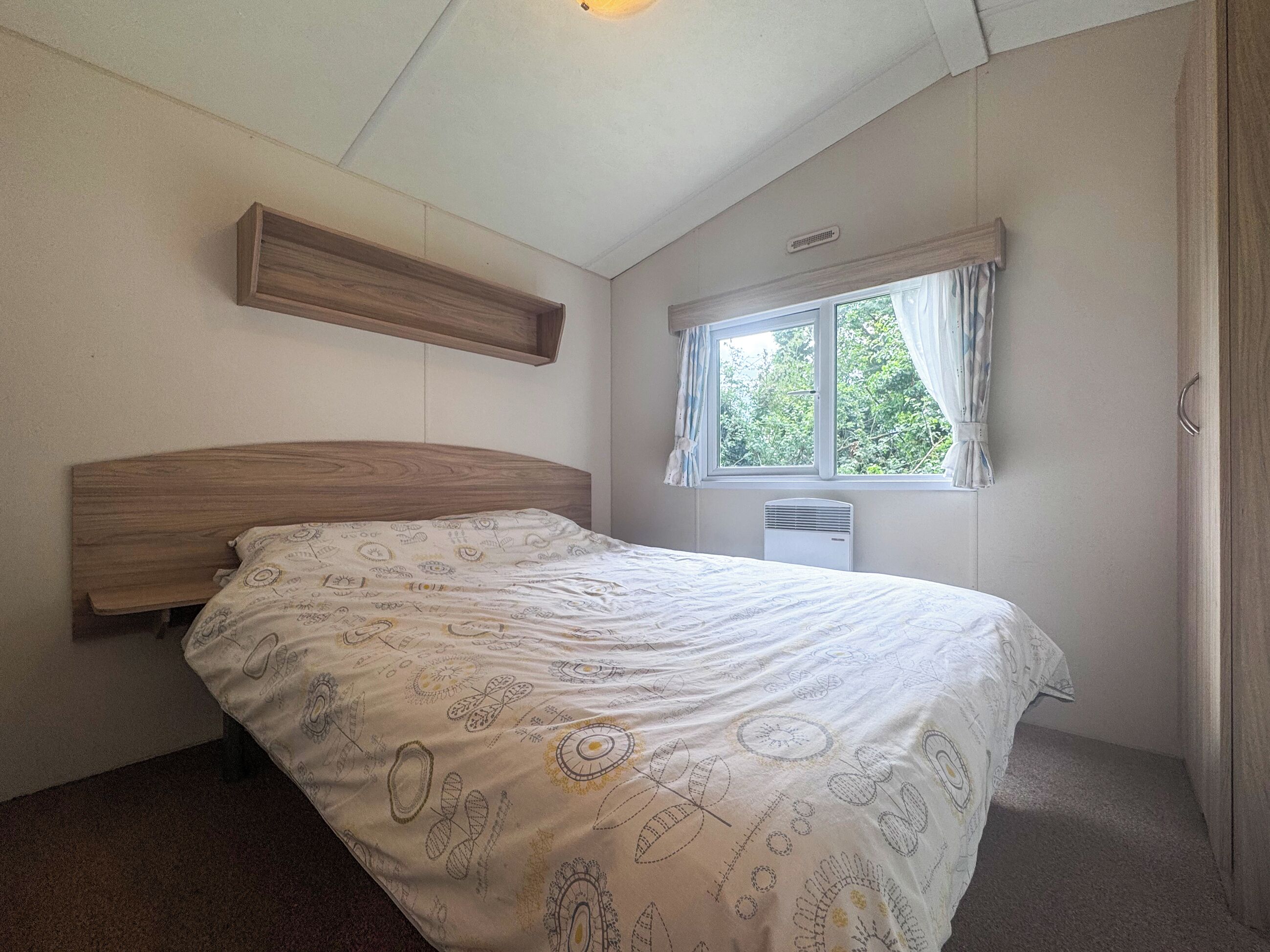 A bedroom with a wooden headboard, a window with curtains, a radiator, and a patterned bedspread.