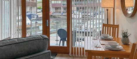 Una habitación con una mesa de comedor preparada para dos personas, un sofá, una silla y vistas a un puerto deportivo con barcos.