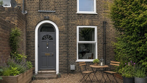A brick house with a grey door, a small garden with a wooden bench and table, and a large tree. - 2 bedroom accommodation in Norwich (Norwich)