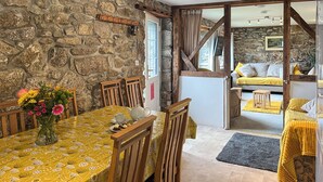A dining area with a stone wall, wooden chairs, a table with a yellow patterned tablecloth, and a vase of flowers. - Mum's Cottage - UK52136 (Minions)