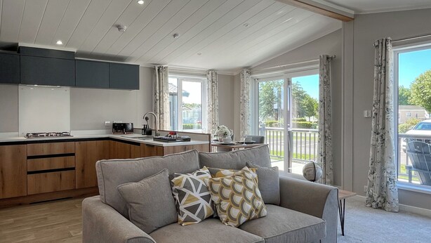 A modern living room with a grey sofa, a kitchen area with wooden cabinets, and large windows offering a view of the outdoors. - Victory Wisteria Lodge (Patrington Haven)