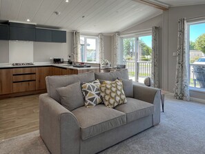 A modern living room with a grey sofa, a kitchen area with wooden cabinets, and large windows offering a view of the outdoors. - Victory Wisteria Lodge (Patrington Haven)