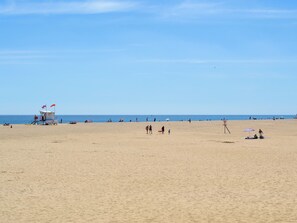 Uma praia com uma torre salva-vidas, céu claro e pessoas ao longe.