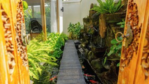 Un estrecho sendero de madera que conduce a un estanque con plantas acuáticas y una pequeña cascada, rodeado de abundante vegetación.