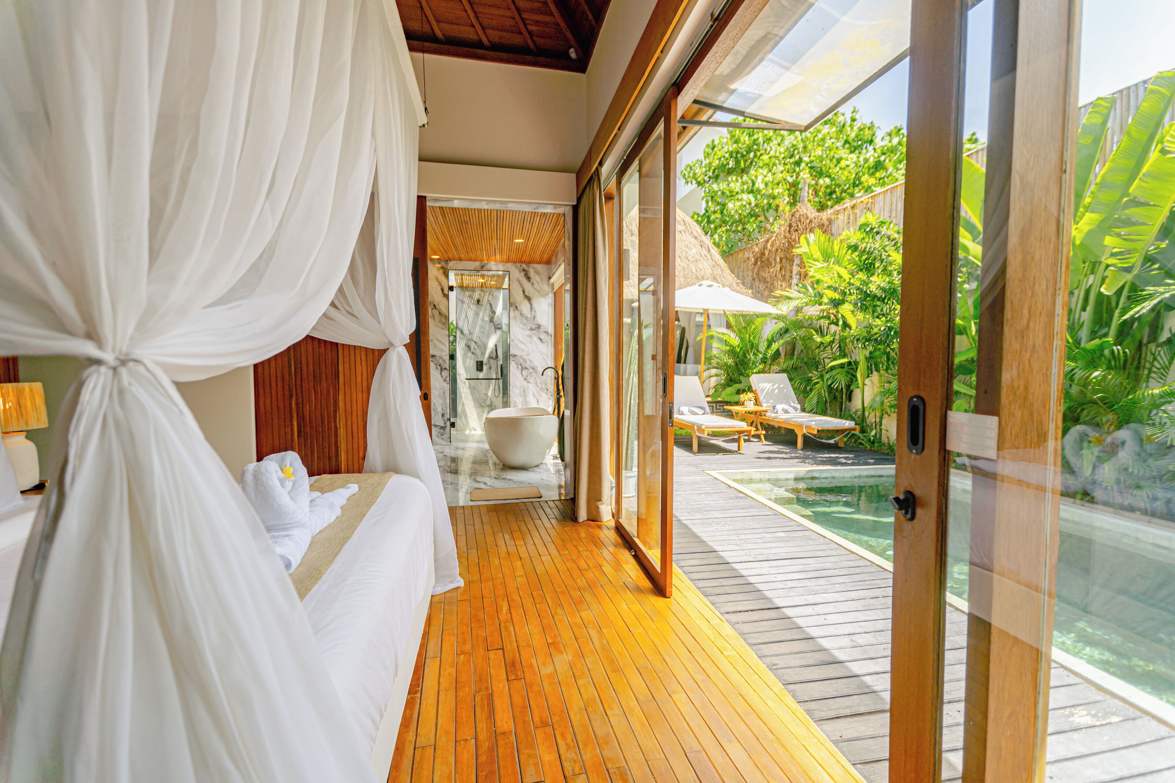 Une chambre avec un plancher en bois, un grand lit avec du linge de lit blanc, une douche en verre et une vue sur une piscine et un espace extérieur.
