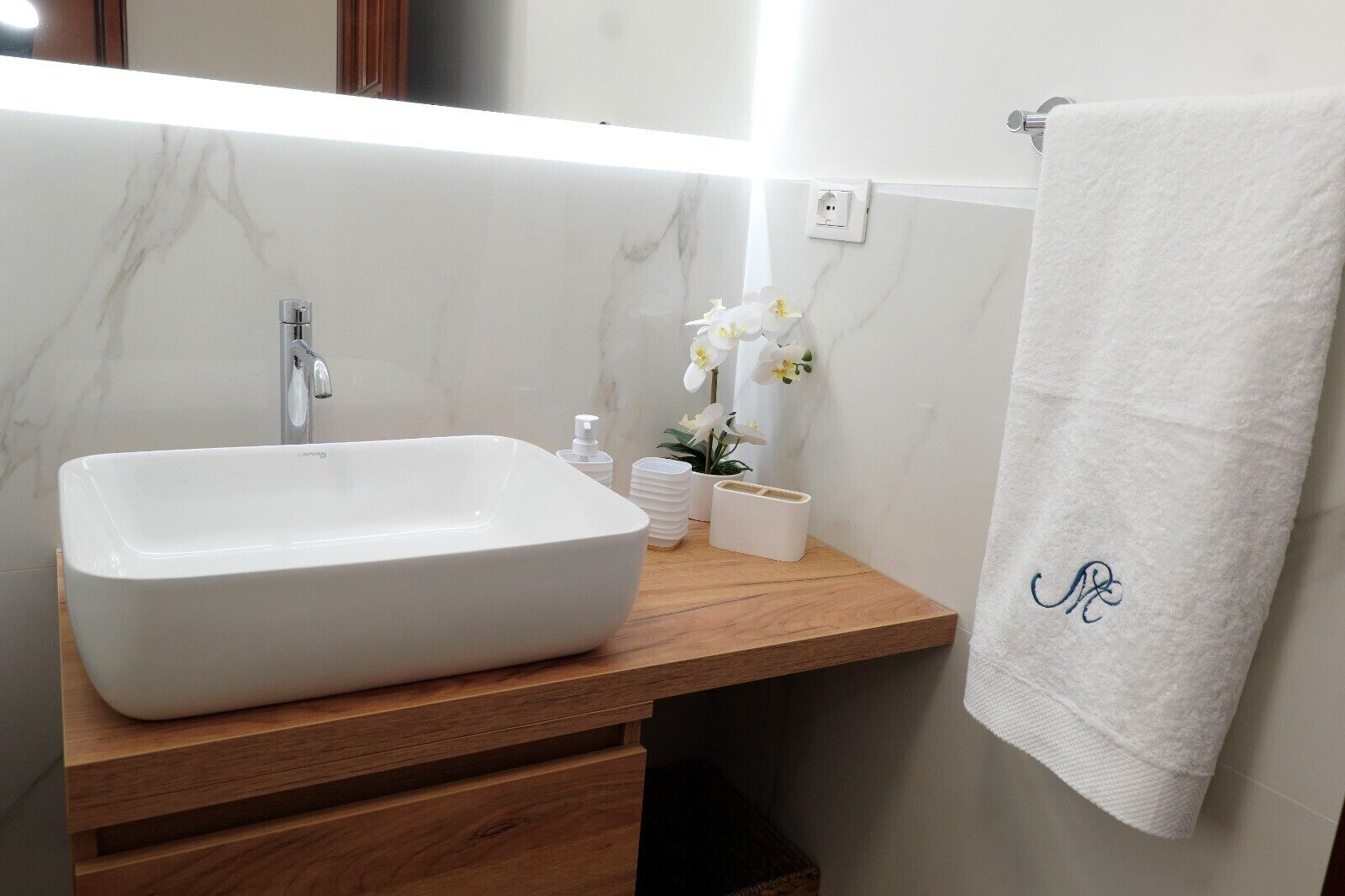 A bathroom with a white sink, a wooden vanity, a white towel with a monogram, and a small orchid plant.