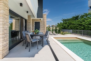 Ein moderner Außenbereich mit Essplatz, Pool, Steinmauern und einer Glastür, die zu einer überdachten Terrasse führt.