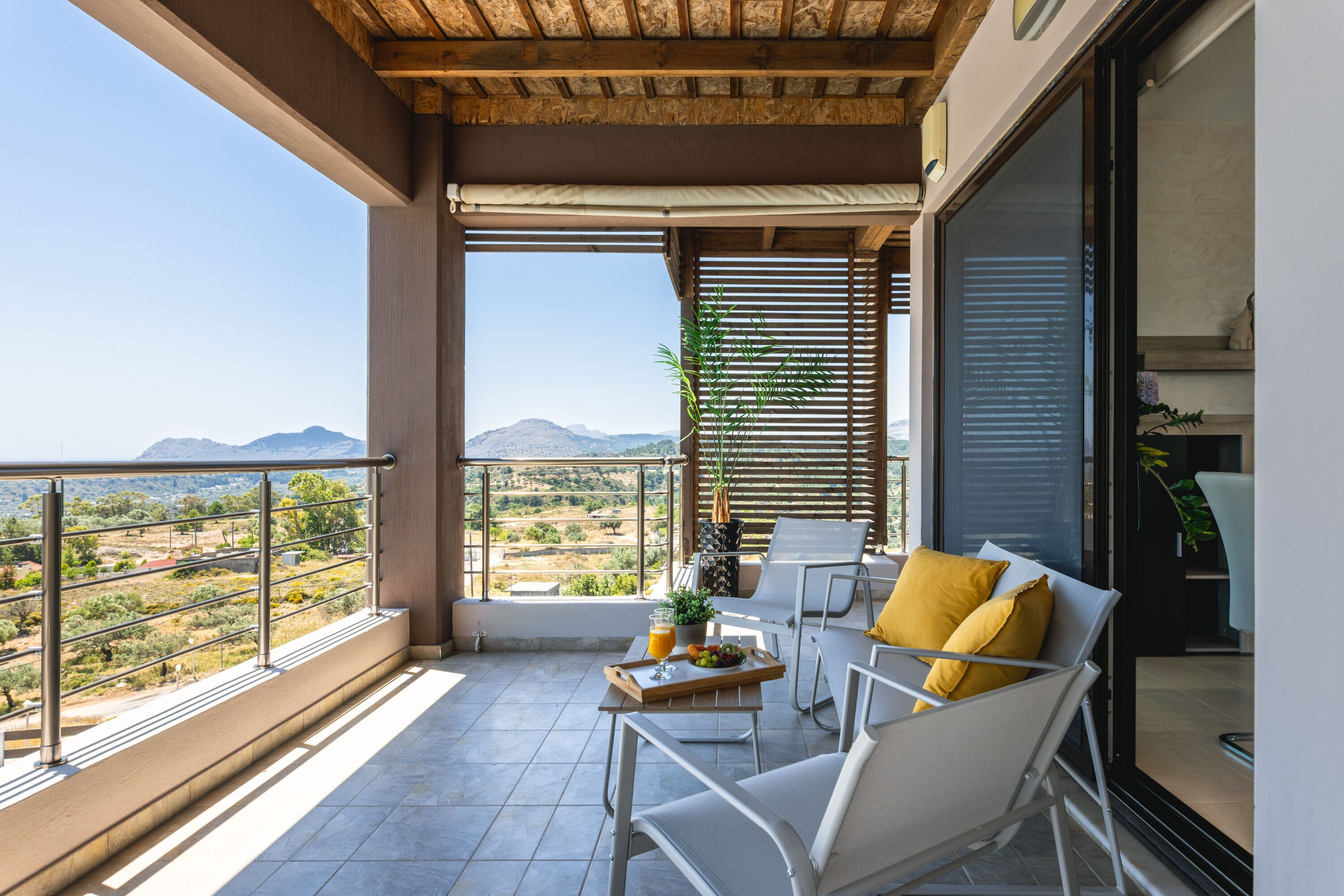 Un balcon avec vue sur les montagnes, une table avec un plateau, et deux chaises blanches dotées de coussins jaunes.