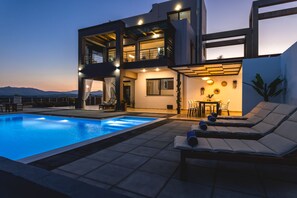A modern two-story house with a swimming pool, outdoor seating, and a view of the mountains.