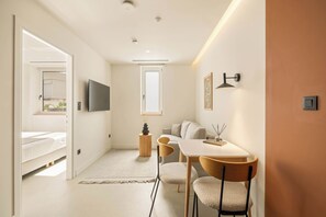 Una camera moderna e luminosa con un letto, una TV, un'area pranzo con sedie e una finestra che lascia entrare luce naturale.