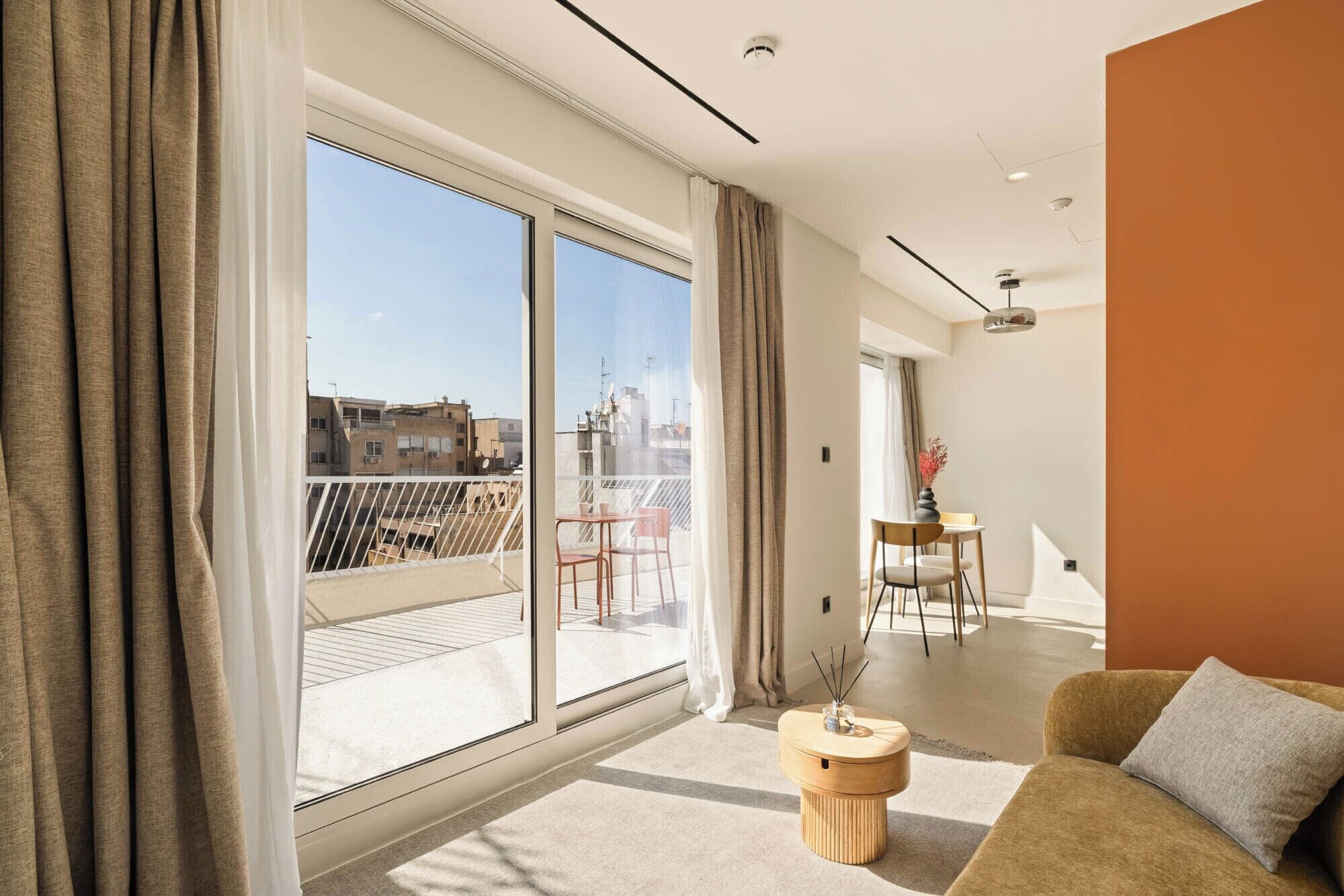 A modern living room with a large sliding glass door leading to a balcony, a small round wooden table, and a sofa with a cushion.