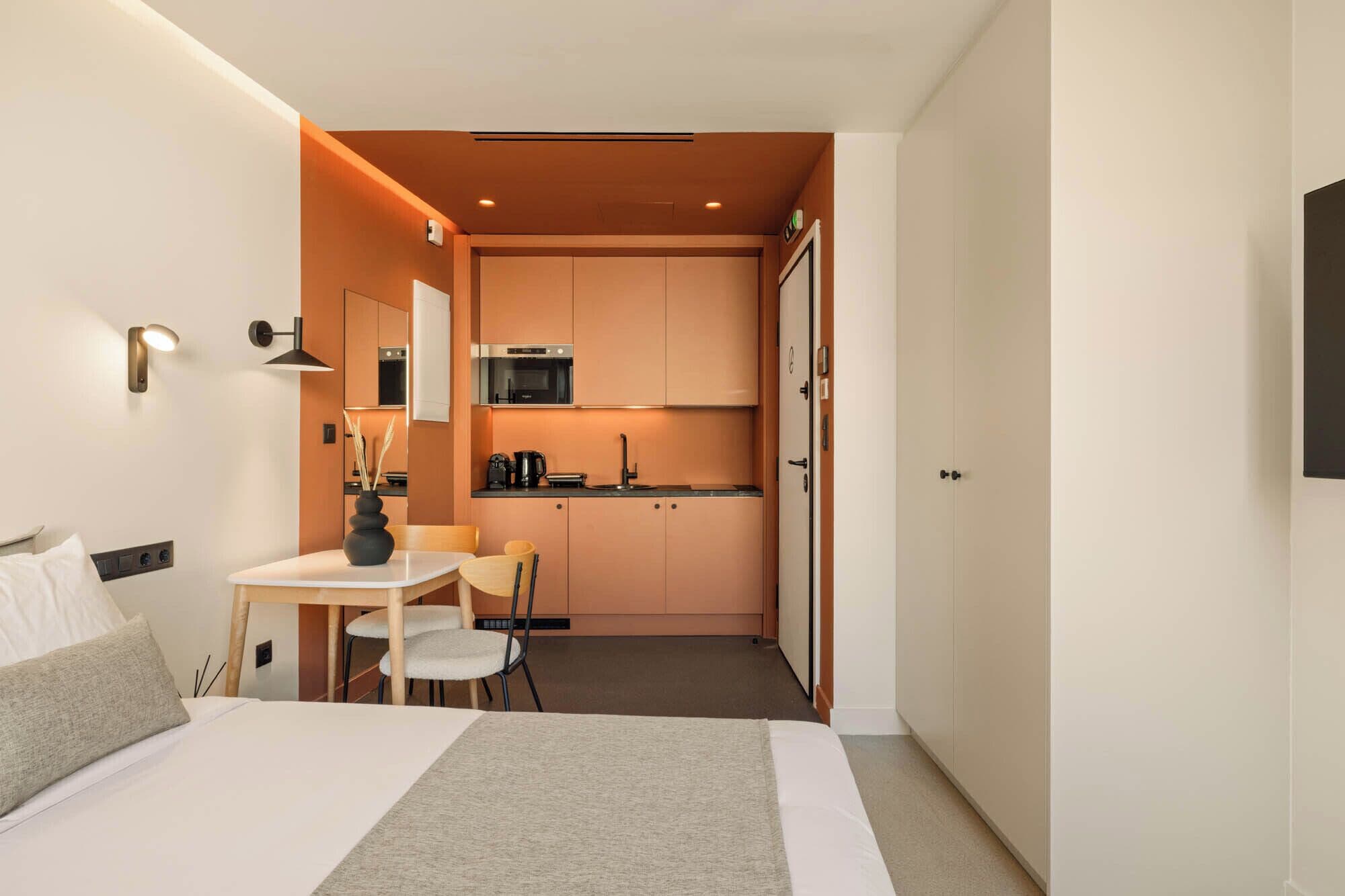 Ein kleines Hotelzimmer mit Bett, Nachttisch und einer kompakten Küchenzeile inklusive Mikrowelle und Kaffeemaschine.