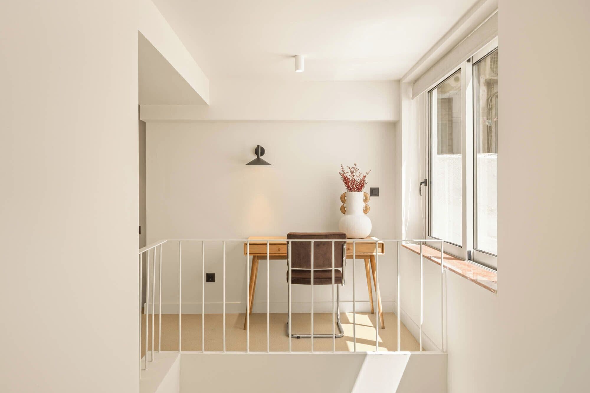 A minimalist room with a wooden table, a chair, a vase with dried plants, and a window with blinds.