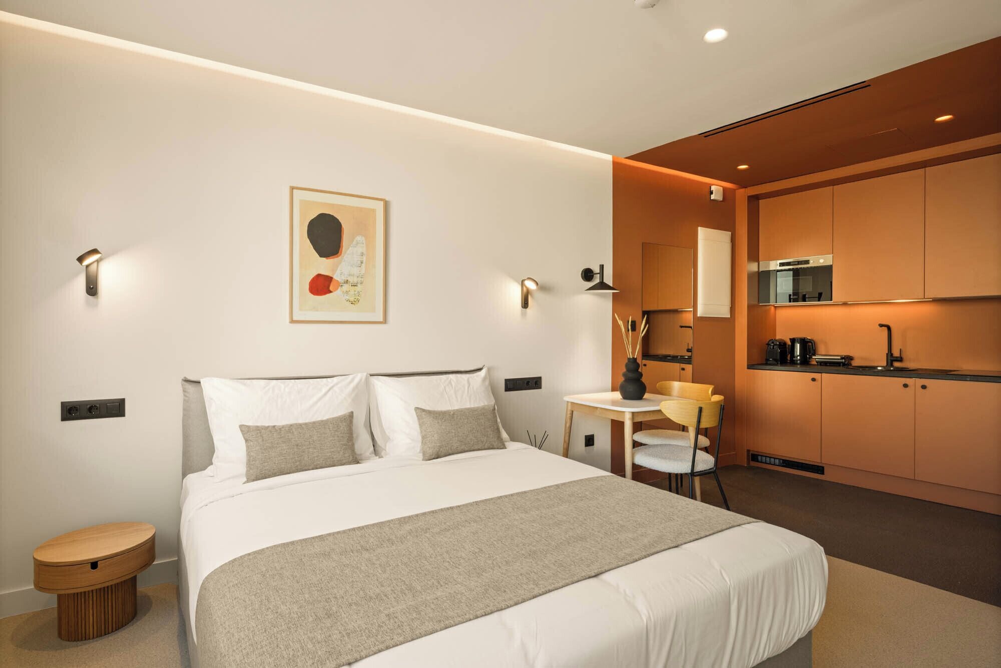 Una habitación de hotel moderna con una cama grande, mesitas de noche, un escritorio pequeño y una kitchenette.