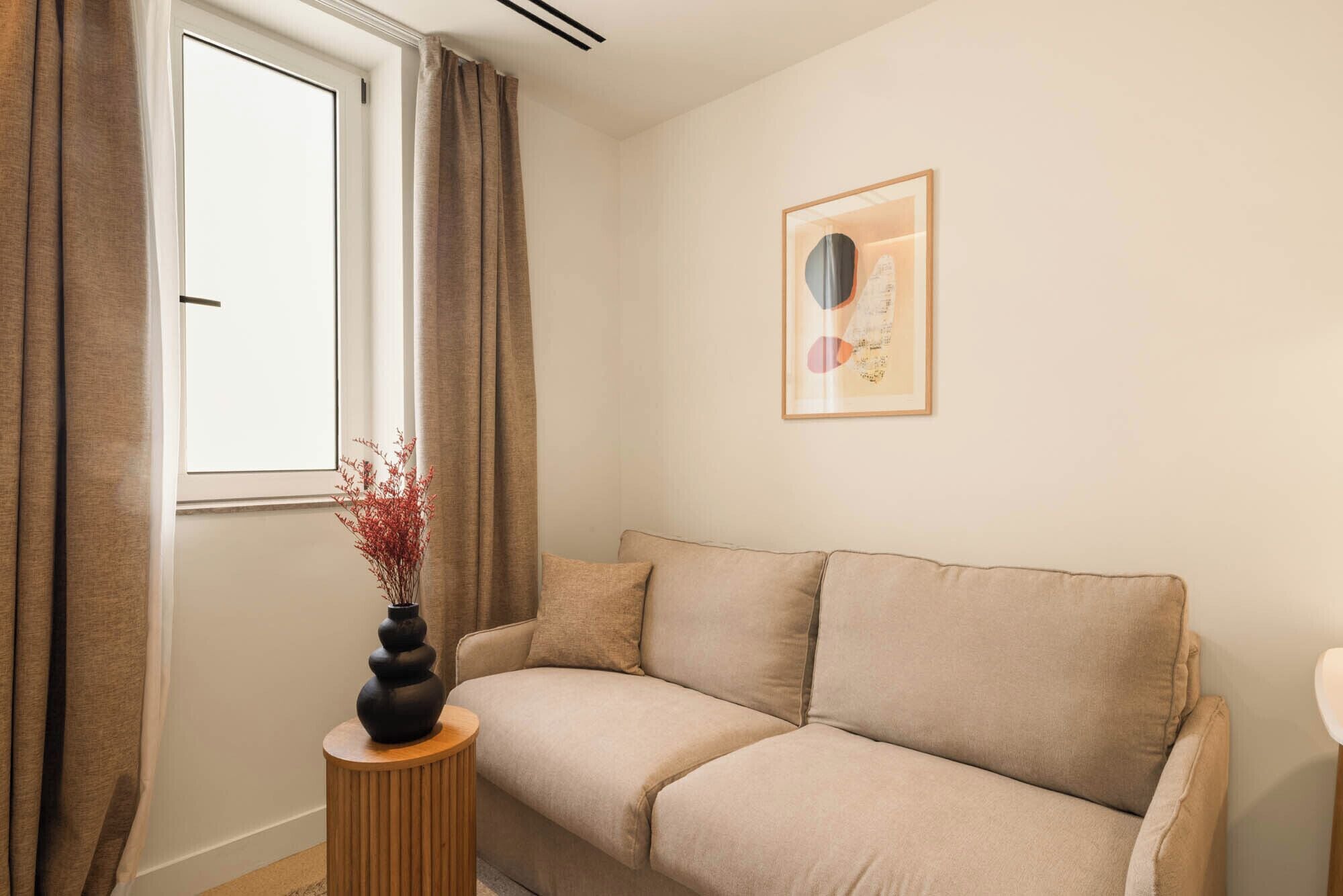 A living room with a beige sofa, a round wooden side table, a vase with red branches, a window with curtains, and a framed abstract artwork on the wall.