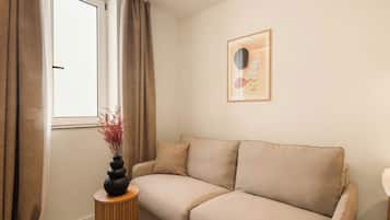Een woonkamer met een beige bank, een rond houten bijzettafeltje, een vaas met rode takken, een raam met gordijnen, en een abstract kunstwerk in een lijst aan de muur.