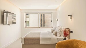 A bedroom with a bed, a TV mounted on the wall, a window with blinds, and a chair. - Grid II by Zoia Living (Athens)