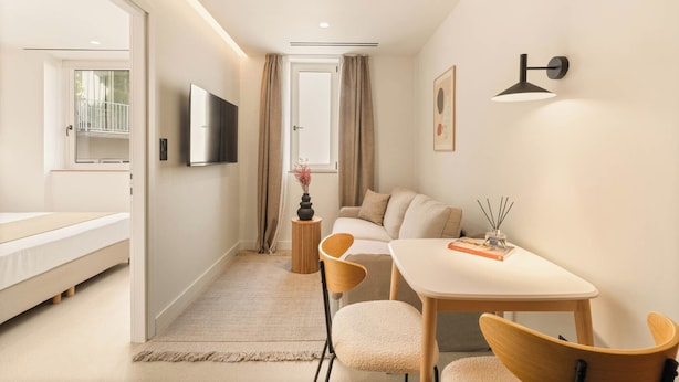 Habitación de hotel moderna con cama, televisión, mesa de comedor con sillas, una mesita auxiliar y una ventana con cortinas.