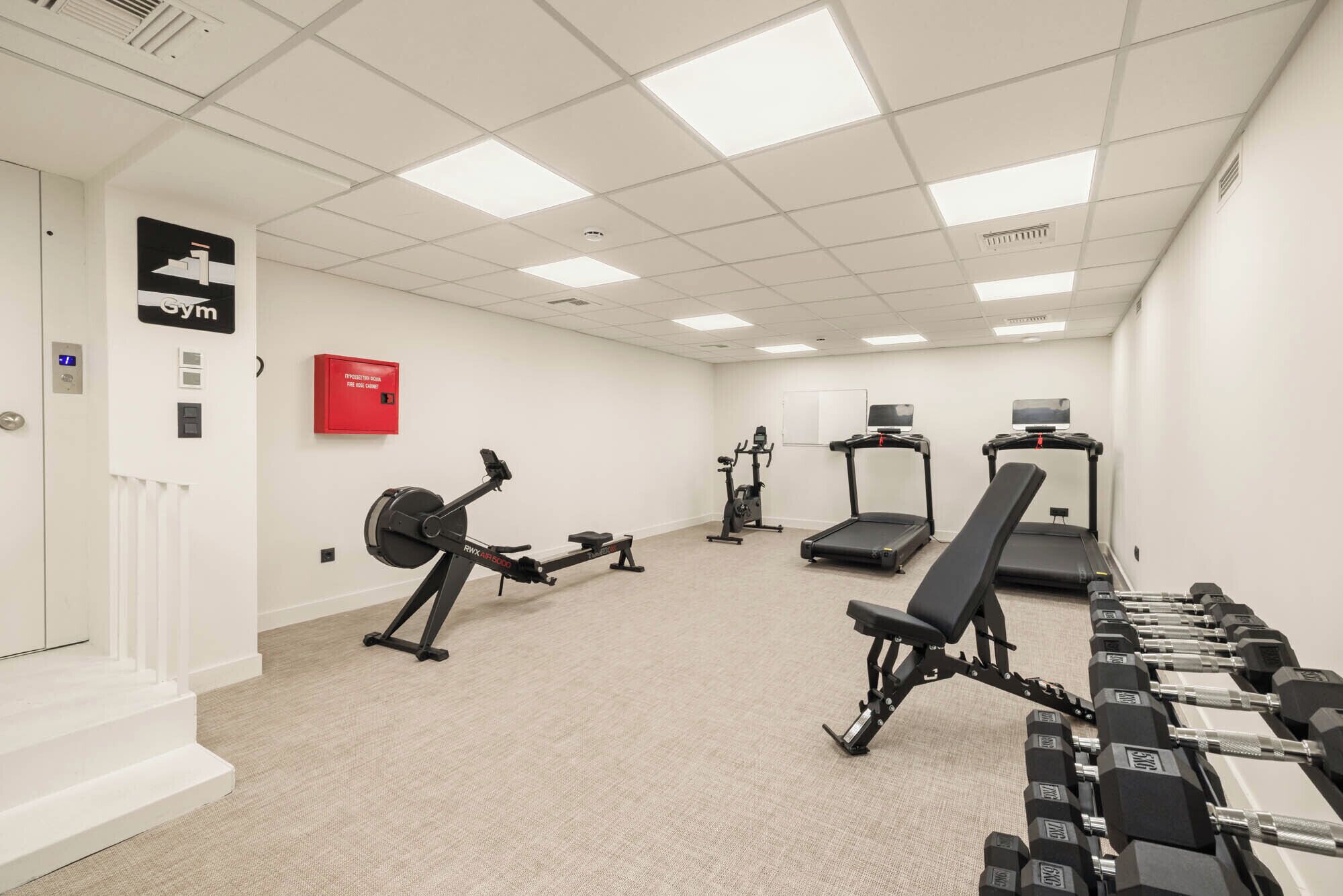 Un gimnasio con equipo cardiovascular, mancuernas y un armario rojo para mangueras contra incendios.