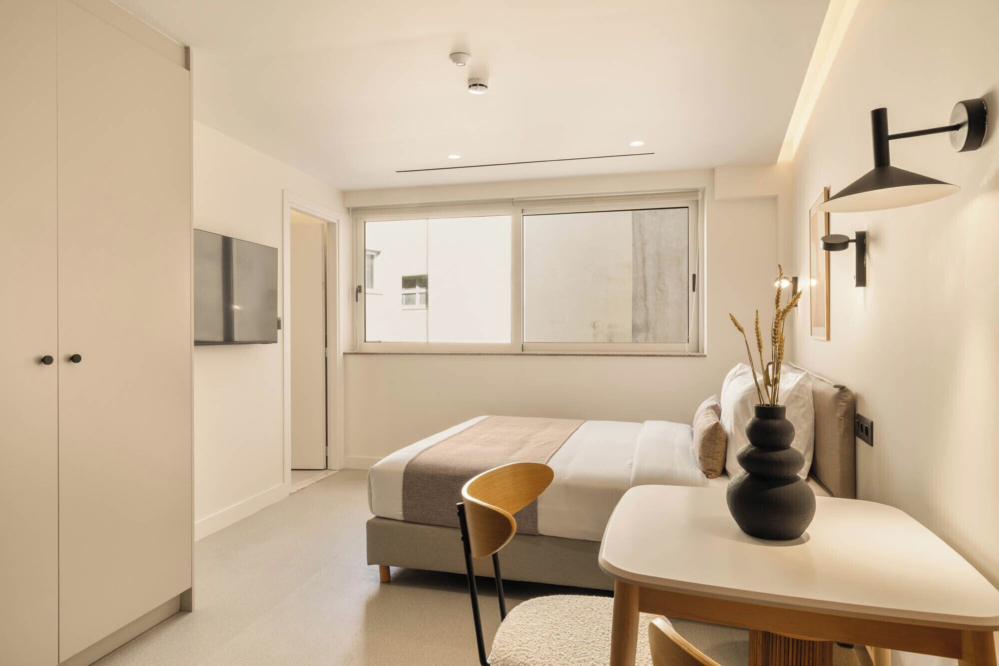 A modern bedroom with a bed, a bedside table, a chair, a TV, and a window.