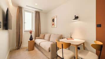 A compact living space with a sofa, a small table, two chairs, a TV mounted on the wall, and a window with curtains.