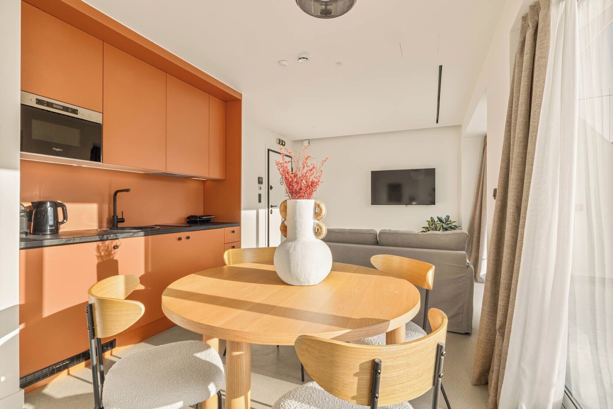 A modern kitchen with orange cabinets, a dining table with chairs, and a living area with a sofa and TV.