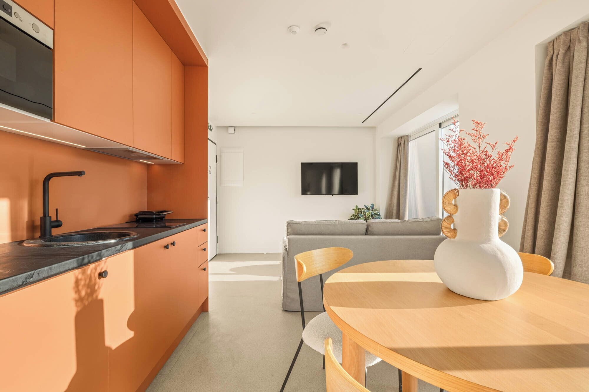 A modern kitchen with a black faucet, a flat-screen TV mounted on the wall, and a dining area with a round wooden table and chairs.