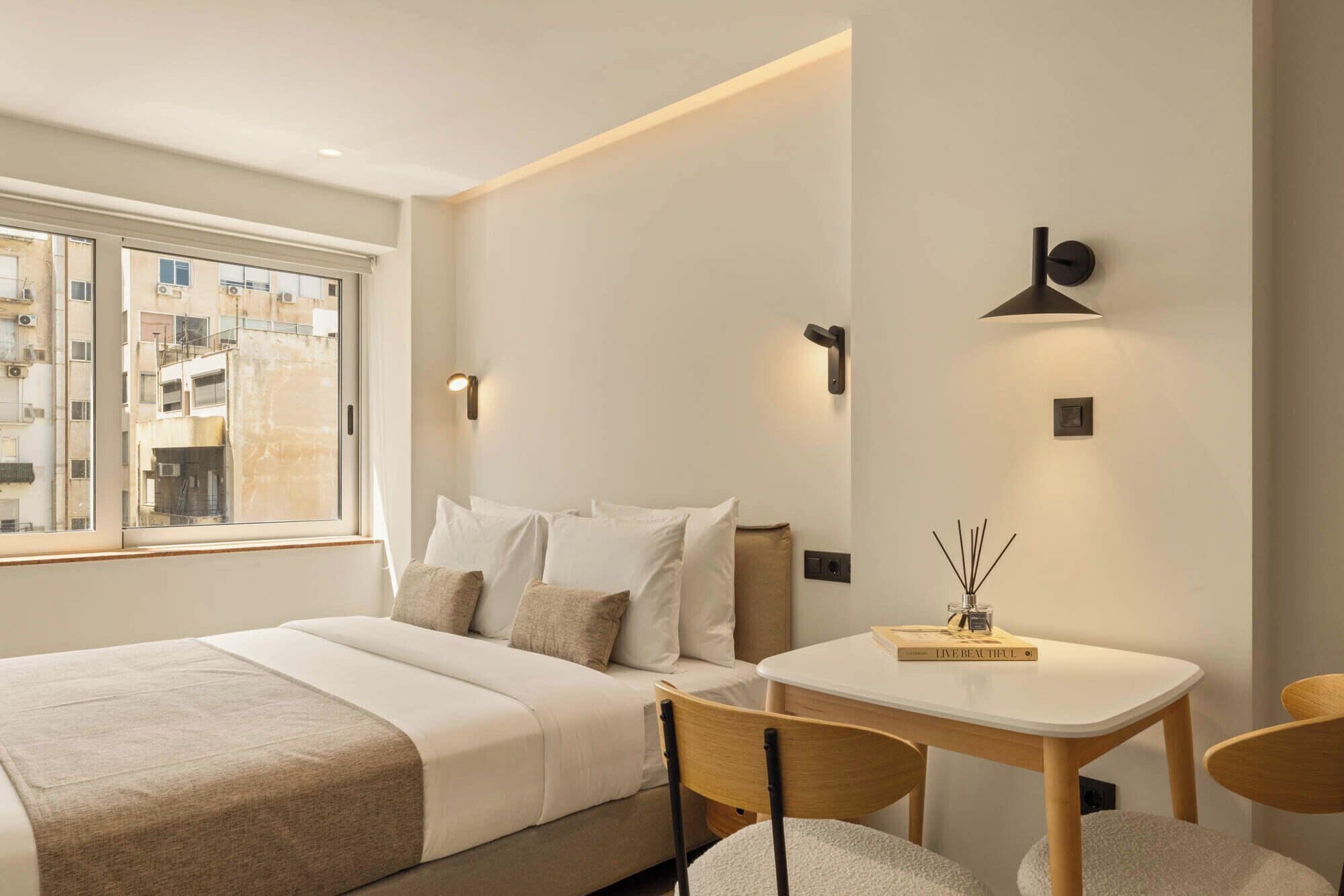 A hotel room with a bed, a small table, two chairs, a window, and wall-mounted lighting.
