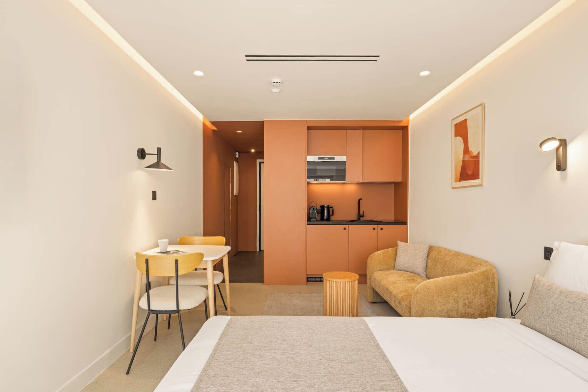 A modern hotel room with a bed, a sofa, a dining area with chairs, a kitchen with cabinets, and a small round table.