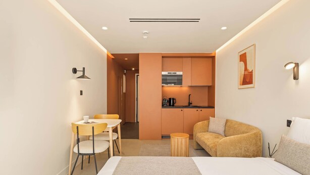 A modern hotel room with a bed, a sofa, a dining area with chairs, a kitchen with cabinets, and a small round table. - Grid by Zoia Living (Athens)