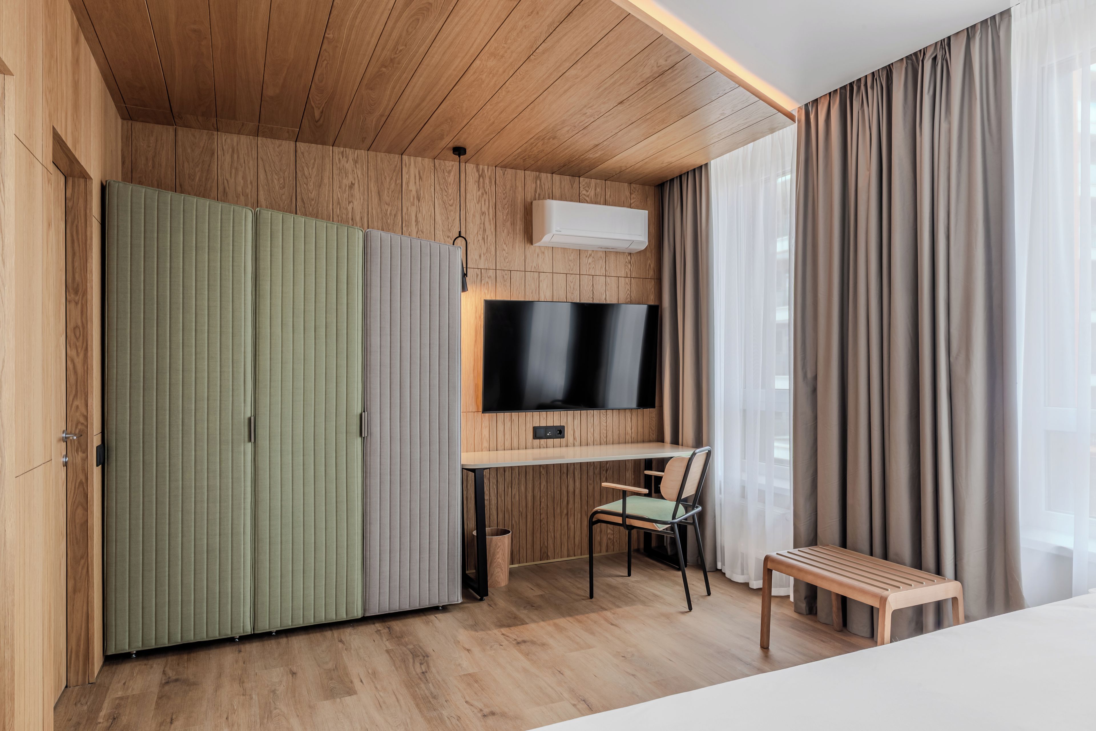 Een moderne kamer met een houten plafond, een groene garderobe, een flatscreen televisie aan de muur, een bureau met een stoel en een raam met gordijnen.