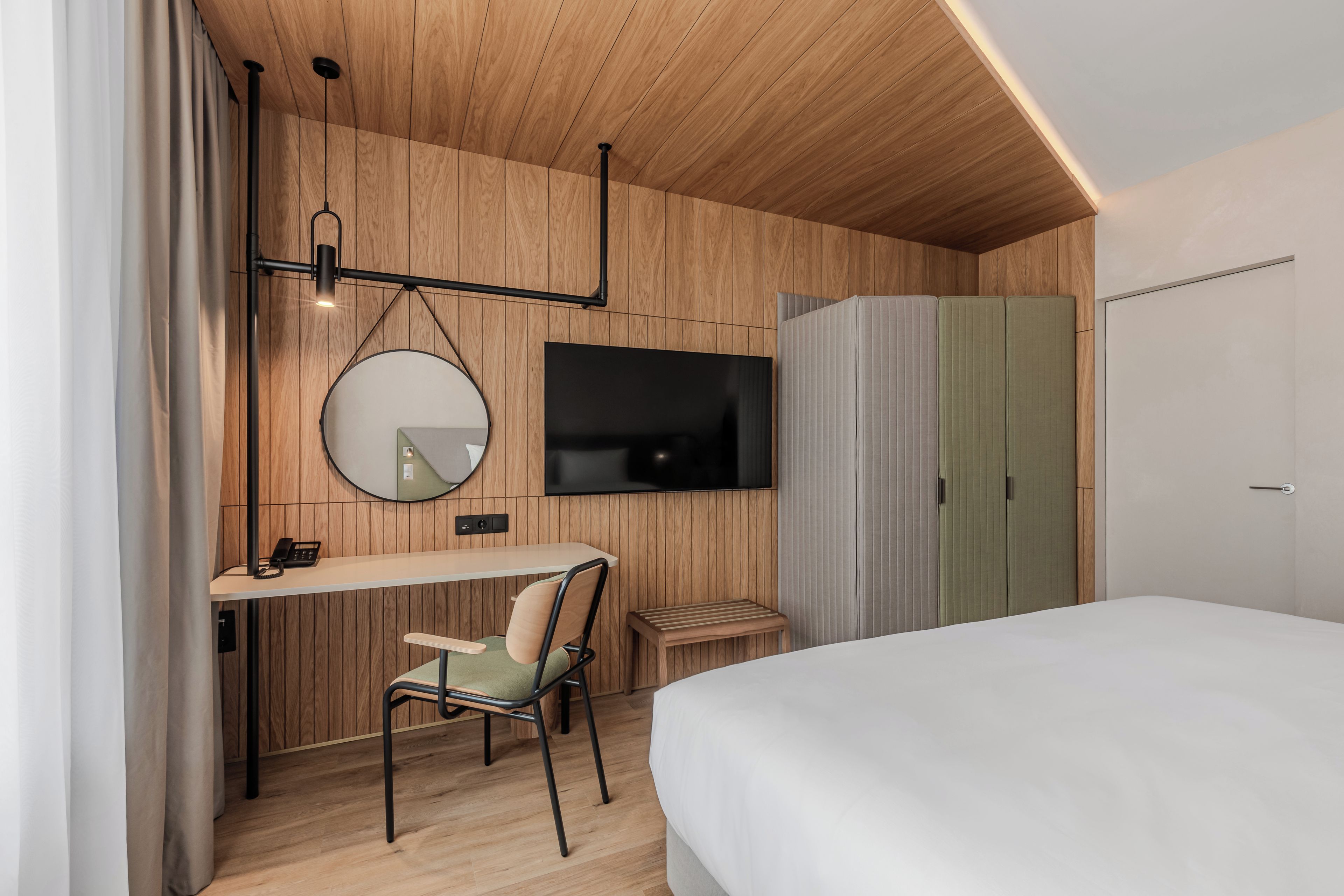 A modern bedroom with a wooden ceiling, a flat-screen TV mounted on the wall, a desk with a chair, and a wardrobe with sliding doors.
