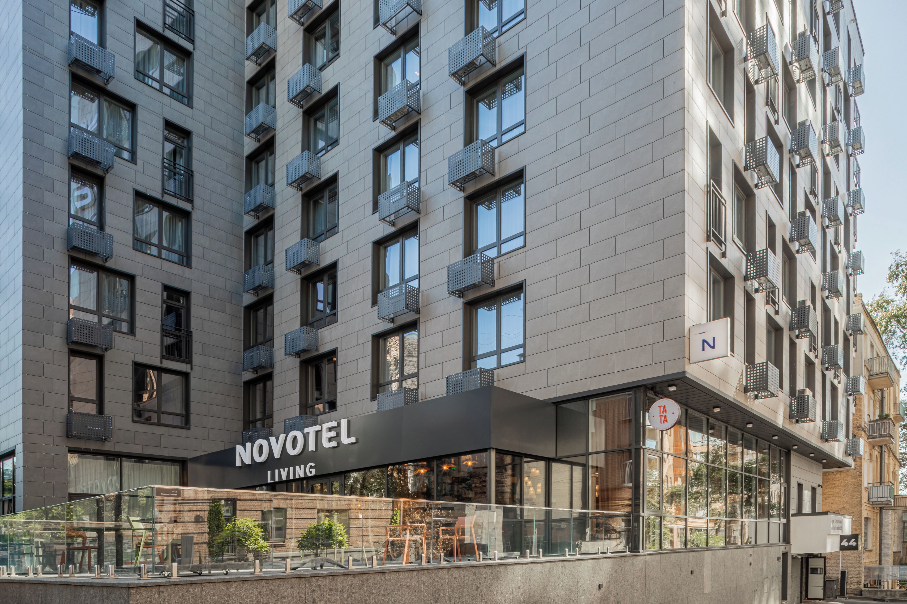 A modern hotel with a prominent 'NOVOTEL LIVING' sign, large windows, and a balcony area with outdoor furniture.