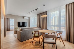 A modern living room with a sofa, a TV, a dining table with chairs, and large windows with curtains. - Novotel Living Kyiv (Kyiv)