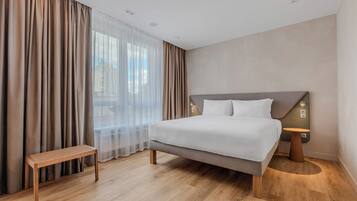 A modern hotel room with a large bed, a bench, and a view of the city through sheer curtains.