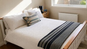 Ein ordentlich bezogenes Bett mit weißer Daunendecke, einer gestreiften Tagesdecke und einem karierten Kopfkissen. Ein Nachttisch mit Lampe und Bilderrahmen ist ebenfalls vorhanden.
