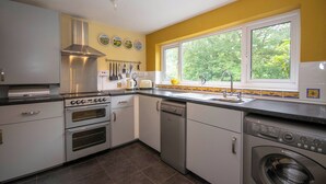 A kitchen with built-in appliances, a window with a view of greenery, and a tiled backsplash. - Railway & Riverside Retreat (Llangollen)