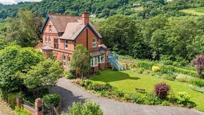 A large brick house with a blue staircase, surrounded by lush greenery and a mountain backdrop. - Railway & Riverside Retreat (Llangollen)