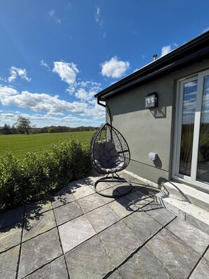 Une terrasse avec un fauteuil suspendu, un sol carrelé en pierre et un bâtiment doté d’une fenêtre et d’une applique murale.