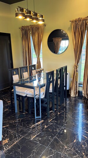Un coin repas avec une table en verre, des chaises foncées, un lustre moderne et un grand miroir au mur.