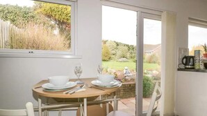 A dining area with a table set for two, a window with a view of a garden, and a coffee machine on the counter. - Bungalow - Sleeps 2 - Parking (Newquay)