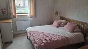 A bedroom with a bed, a bedside table, a window with curtains, a radiator, and a hanging light fixture. - Golspie, Firthview, A Cosy Seafront Escape (Golspie)