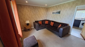 A living room with a dark brown sectional sofa, a small wooden side table with a lamp, and a single chair. - Golspie, Firthview, A Cosy Seafront Escape (Golspie)