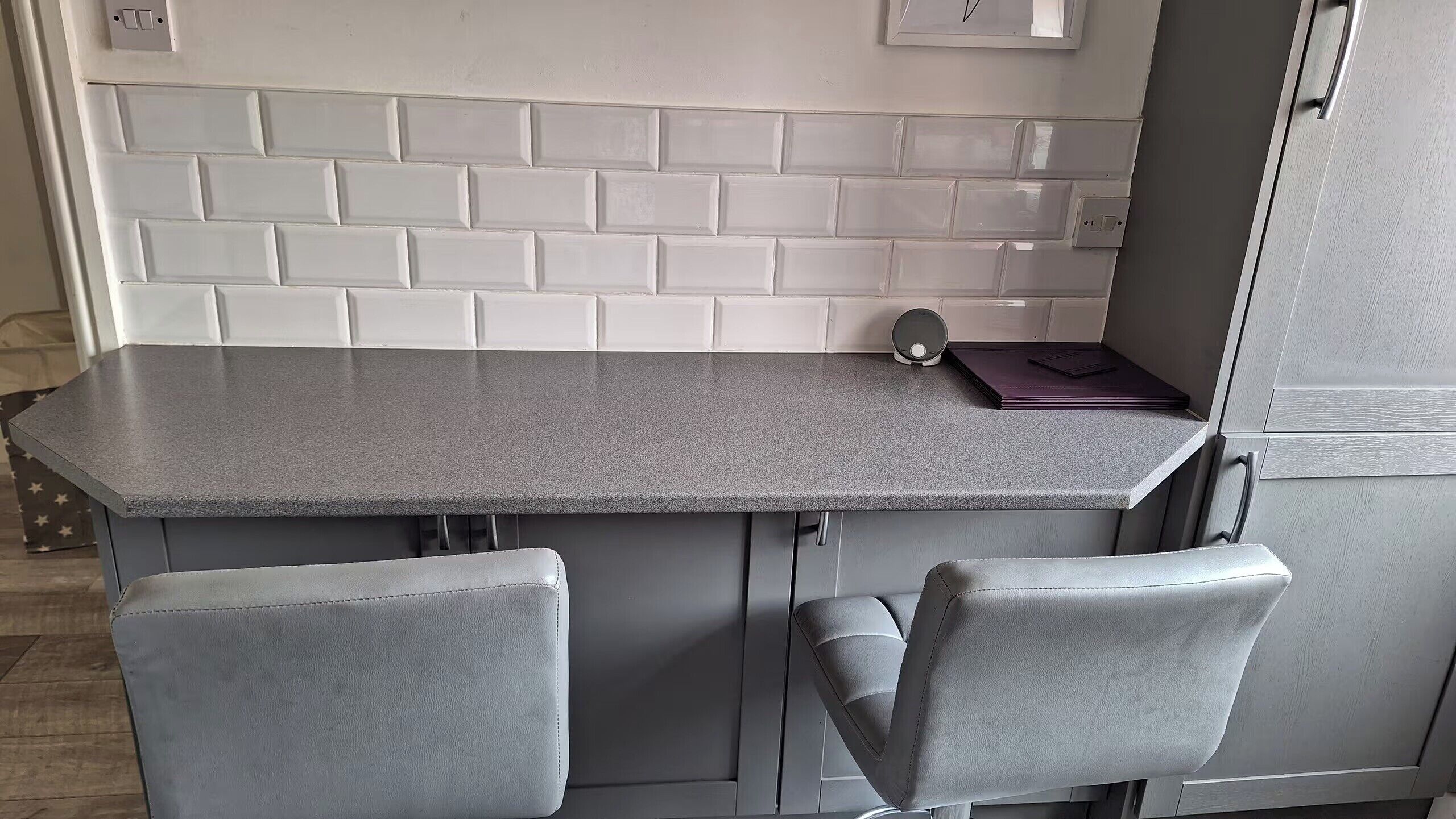 A kitchen with a grey countertop, white tiled backsplash, and two grey chairs.