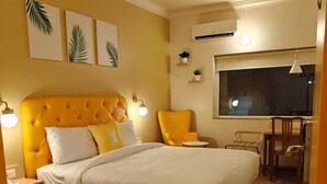 Un dormitorio con una cama grande, una mesita de noche, una silla, una pequeña mesa y una ventana con aire acondicionado.
