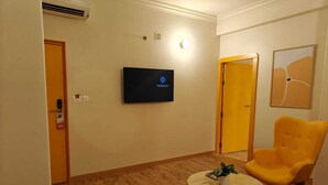 Una habitación de hotel con una televisión montada en la pared, una mesa redonda de centro con una planta, un sillón amarillo y un pequeño reposapiés.