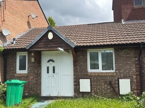 A residential house with a white front door, two windows with shutters, a brick facade, and a green trash bin in the yard. - Bungalow Sleeps 3 With Parking and Garden (Manchester)