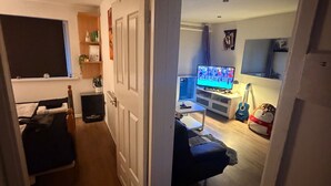 A narrow hallway leading to a living room with a television, a guitar, and a blue sofa.