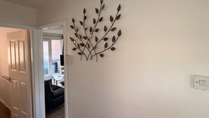 A hallway with a wall-mounted decorative metal leaf art piece, a white door, and a glimpse of a living area with a sofa and a table.